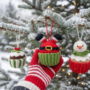以下が含まれることがあります： 雪の降る常緑樹の枝に吊るされた、3つのかぎ針編みのクリスマスオーナメント。オーナメントはカップケーキの形で、サンタクロース、雪だるま、ヒイラギベリーのカップケーキが含まれています。サンタのオーナメントは赤い体、黒いブーツ、金のバックルが付いています。
