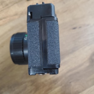 May include: A black and silver film camera with a textured grip. The camera is in a folded position, showing the lens and the top of the camera.