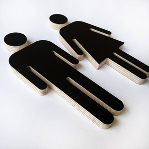 May include: Two black wooden figures, one male and one female, are cut out in a layered style. The figures are standing side by side.