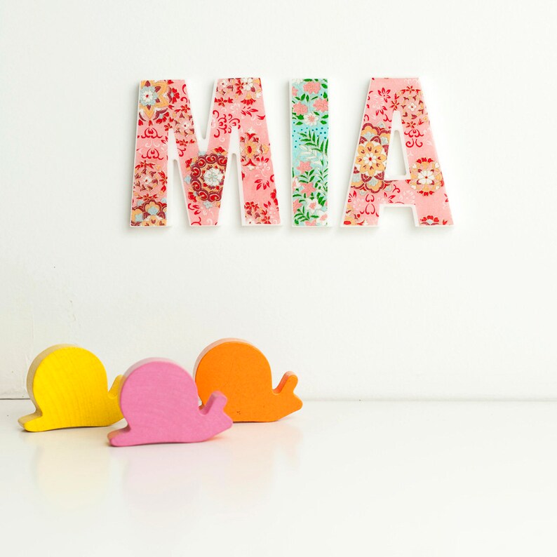 Peut inclure: Lettres en bois orthographiant le nom "MIA" en rose, blanc et bleu avec un motif floral. Trois escargots en bois jaune, rose et orange sont au premier plan.