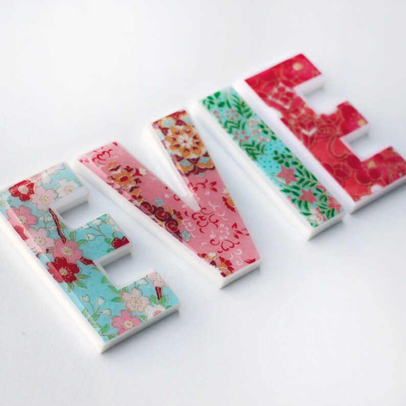 Peut inclure: Le nom "EVIE" &eacute;crit en lettres color&eacute;es et &agrave; motifs. Chaque lettre est en plastique blanc avec un motif floral dans des tons de rose, rouge, bleu et vert.