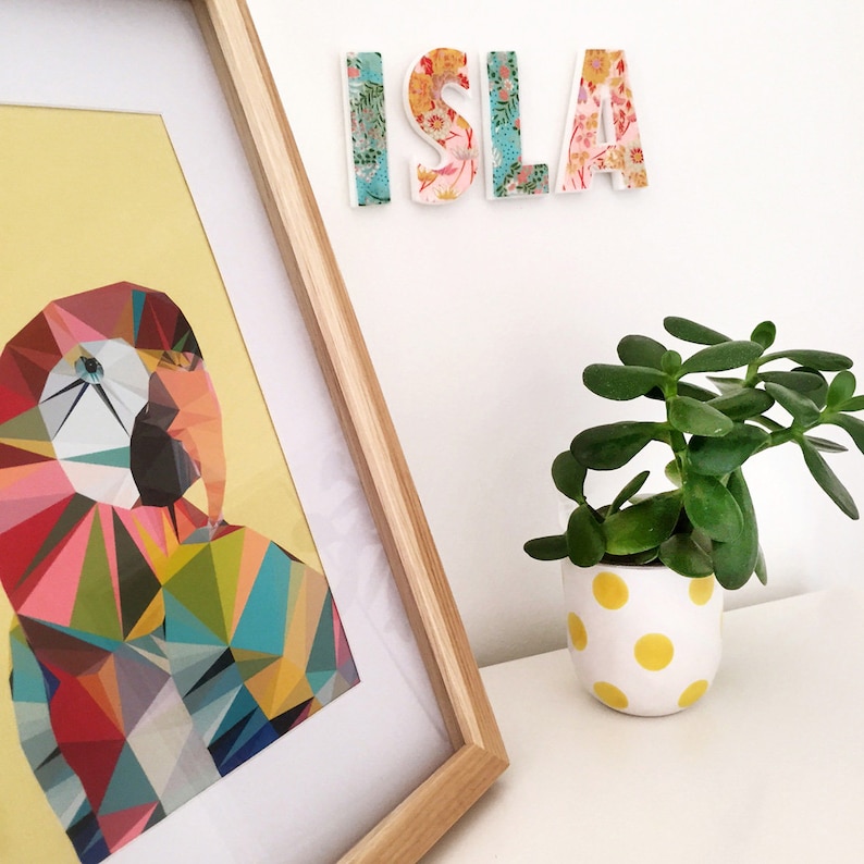 Peut inclure: Une impression encadr&eacute;e d'un perroquet g&eacute;om&eacute;trique color&eacute; sur fond jaune. L'impression est accroch&eacute;e &agrave; un mur blanc avec une pancarte de nom &agrave; motifs floraux qui lit "ISLA" au-dessus. Une petite plante en pot avec des feuilles vertes est sur une surface blanche sous l'impression.