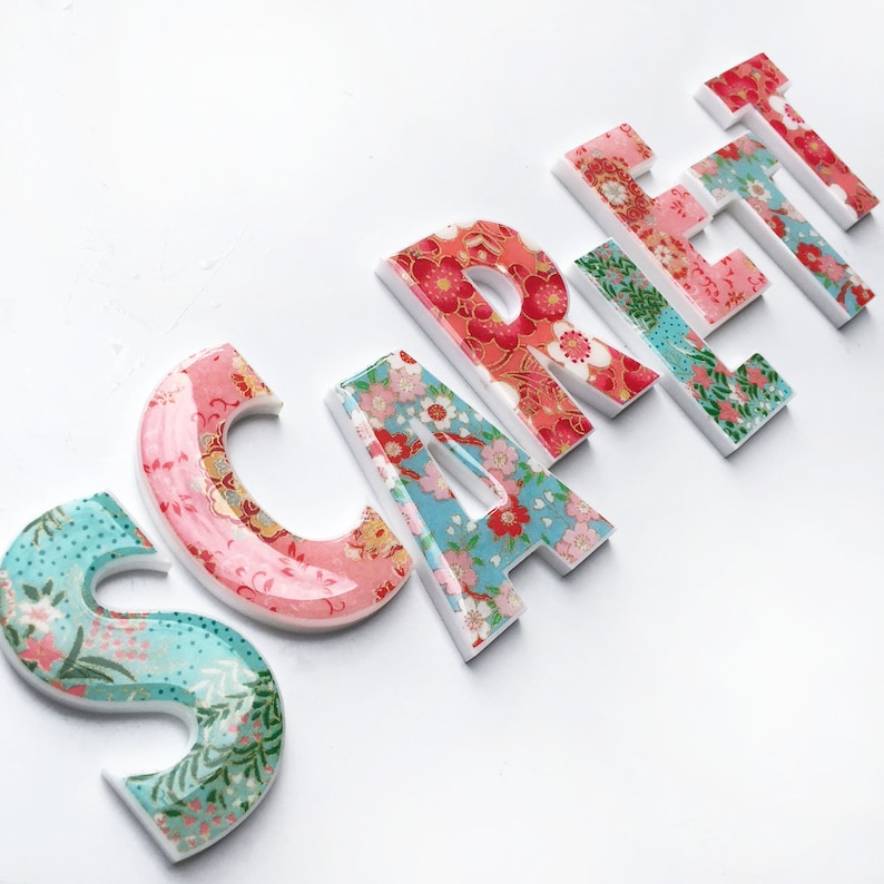 Peut inclure: Le nom "SCARLETT" &eacute;crit en lettres blanches avec un motif floral. Chaque lettre est d&eacute;cor&eacute;e d'un motif floral diff&eacute;rent dans des tons de rose, rouge, bleu et vert.