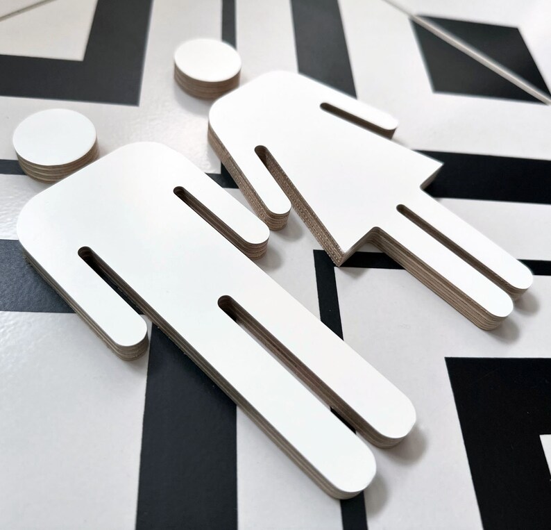 May include: Two white wooden cutout figures, one with a skirt and the other with pants, are displayed on a black and white patterned surface.