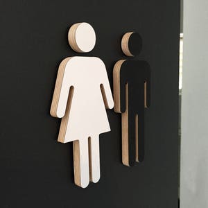 May include: Two wooden cutout figures, one white and one black, representing the male and female gender symbols. The figures are mounted on a black background.