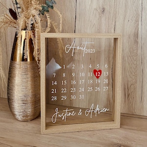 Peut inclure: Un cadre en bois rectangulaire contenant une vitre transparente imprimée avec le calendrier du mois d'Aout 2023. Le calendrier présente des chiffres blancs, un cœur rouge sur le 12 et les prénoms Justine & Adrien en écriture blanche.