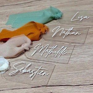 Peut inclure: Étiquettes de nom en acrylique transparent avec lettrage blanc en cursive, avec des noms comme Lisa, Nathan et Mathilde. Les étiquettes sont disposées sur une surface en bois, avec des rubans de tissu colorés verts, oranges et beiges.