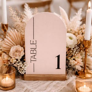 May include: A blush pink table number sign with the word "TABLE" printed vertically and the number "1" in black. The sign is arched at the top and sits on a wooden base. The background includes floral arrangements and candles.