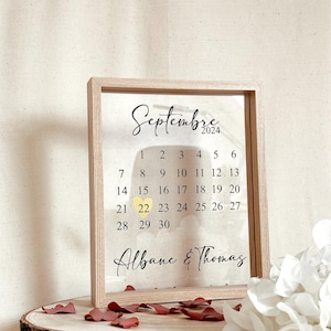 Peut inclure: Calendrier encadré en bois clair pour septembre 2024, avec le 22 mis en évidence. Les noms "Albane & Thomas" sont écrits en dessous. Le calendrier est posé sur une tranche de bois, avec des pétales de rose éparpillés.