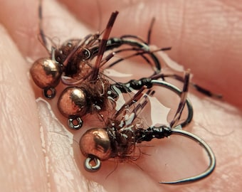 May include: Close-up of three fishing flies. Each fly has a bronze-colored bead head, a black hook, and brown and black fibers. The flies are resting on a wet surface, likely a hand.