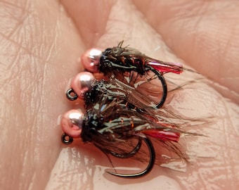 May include: Close-up of three fishing flies. Each fly has a pink bead head, black body, and red accents. The hooks are black and curved. The flies are resting on a light-colored surface.
