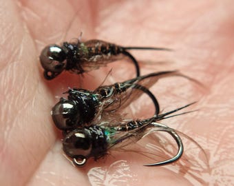 May include: Close-up of three fishing flies. Each fly has a dark, metallic bead head, a black hook, and a body of brown and iridescent materials. The flies are held in a hand, showcasing their intricate details.