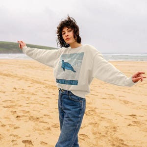 May include: A cream-coloured sweatshirt featuring a blue turtle design and the words "POWER OCEANS". The sweatshirt is worn with blue jeans. The photo was taken on a sandy beach with the sea in the background.