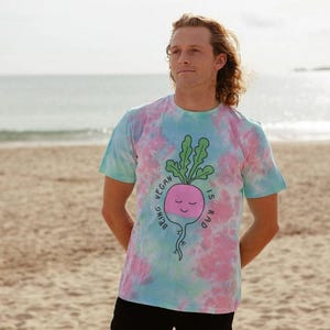 Può includere: Una maglietta tie-dye con un motivo rosa, blu e bianco. La maglietta presenta un ravanello dei cartoni animati con la scritta "Being Vegan is Rad". La maglietta a maniche corte viene indossata su una spiaggia.