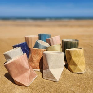 Puede incluir: Una colección de doce vasos de cerámica con superficies geométricas facetadas en varios colores, incluyendo rosa, amarillo, azul, verde, beige y blanco moteado. Cada taza presenta un diseño angular único y un acabado esmaltado suave.