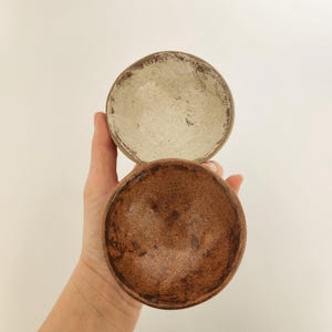 May include: Two handmade ceramic bowls. The top bowl is a light beige colour with a textured surface and a dark brown rim. The bottom bowl is a darker brown colour with a similar textured surface.