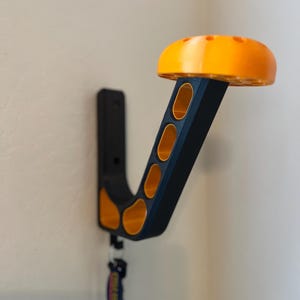 May include: A black and orange wall-mounted hook with a rounded top and multiple holes. The hook is designed to hold items like hats, coats, or bags.