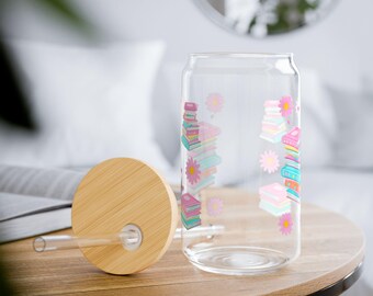 Puede incluir: Un vaso transparente con tapa de bambú y pajita de cristal. El vaso presenta un patrón repetitivo de libros apilados de colores en tonos rosa, azul y turquesa, intercalados con pequeñas ilustraciones de flores rosas en el exterior.