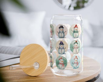 Puede incluir: Un vaso de cristal transparente con tapa de bambú y pajita de cristal. El vaso presenta un patrón repetitivo de muñecos de nieve estilo acuarela en marcos circulares, cada uno con gorros y bufandas de diferentes colores sobre un fondo claro.
