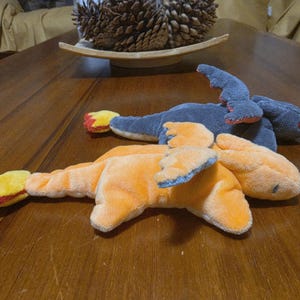 May include: Two plush dragon toys, one orange and one dark grey, lying flat. Each toy features small wings, a long tail ending in a yellow and red flame-like tip, and embroidered eyes. The orange dragon is positioned in the foreground.