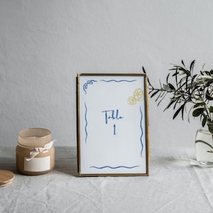 May include: Gold-framed table number card with a white background and blue and yellow illustrations. The card reads "Table 1".