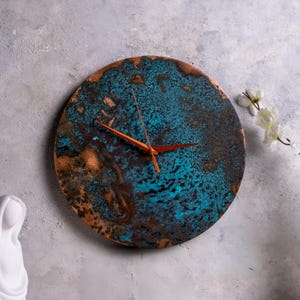 Pode incluir: Relógio de parede redondo com uma superfície texturizada em tons de azul-turquesa, castanho e cobre. Os ponteiros do relógio são de cor laranja brilhante. Um item decorativo para a casa.