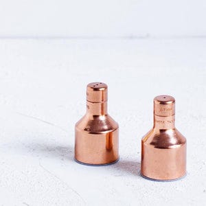 Puede incluir: Un juego de dos saleros y pimenteros de cobre. Los saleros son redondos y tienen un diseño sencillo.