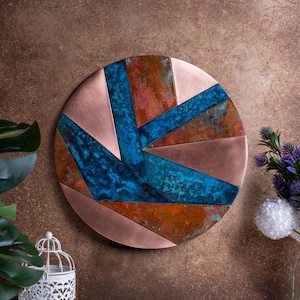 May include: A circular wall art piece made of copper with a geometric design. The copper has been oxidized to create a blue and rust colored patina.