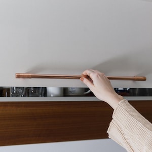 May include: A close-up of a copper cabinet pull handle. The handle is attached to a white cabinet door. A hand is reaching for the handle.