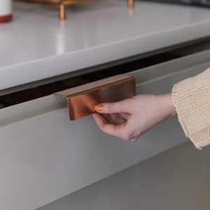 Może przedstawiać: Zbliżenie na miedziany uchwyt szuflady, z ręką sięgającą, aby otworzyć szufladę. Szuflada jest jasnoszara.