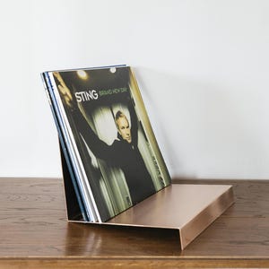 May include: A copper record stand with a stack of vinyl records. The top record is a Sting album titled "Brand New Day".