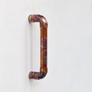 Puede incluir: Una manija de puerta de tubo de cobre con un acabado único e iridiscente. La manija tiene forma de 'D' y está montada en una pared blanca.