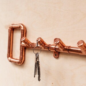 May include: Copper key-shaped wall mounted key holder with four hooks. A set of keys is hanging on one of the hooks.