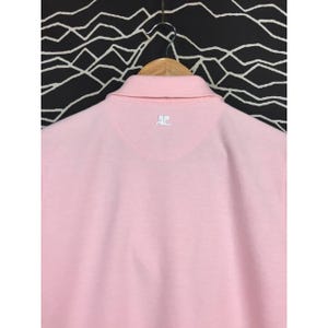 May include: A light pink polo shirt with a collar and short sleeves. The back of the shirt features a small white logo at the neckline. The shirt is hanging on a wooden hanger against a black and white patterned background.
