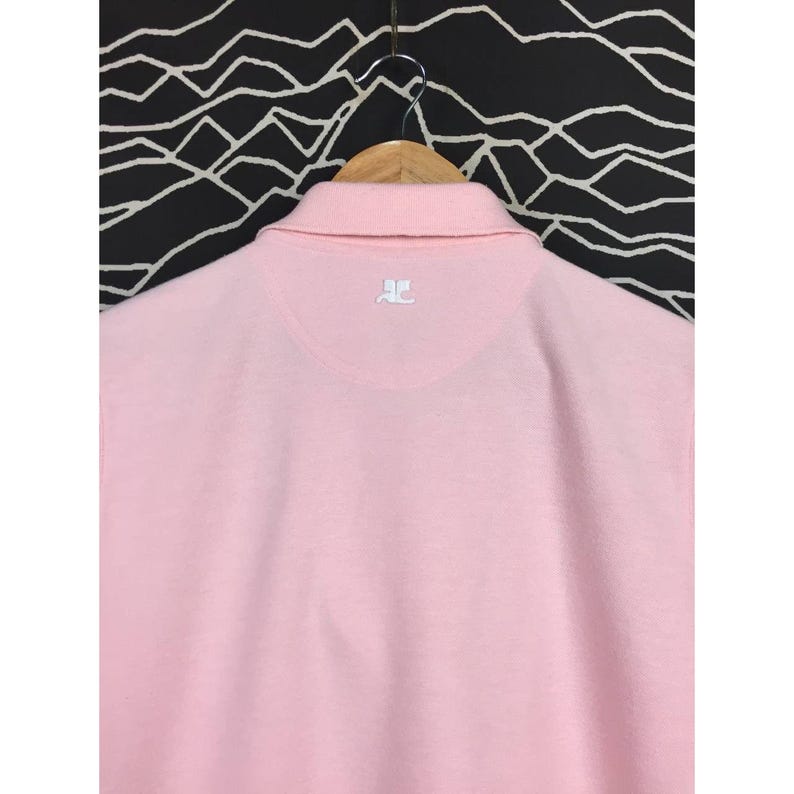 May include: A light pink polo shirt with a collar and short sleeves. The back of the shirt features a small white logo at the neckline. The shirt is hanging on a wooden hanger against a black and white patterned background.