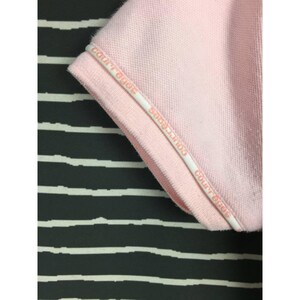 May include: Close-up of a light pink polo shirt sleeve. The sleeve edge has a white trim with the text "COURR&Egrave;GES" in a repeating pattern. The background is a black and white striped pattern.