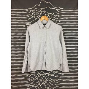 May include: A pale grey long-sleeved shirt with a collar, suspended from a wooden hanger. The shirt features a classic design with a single black button at the collar. The backdrop is a black and white graphic pattern.