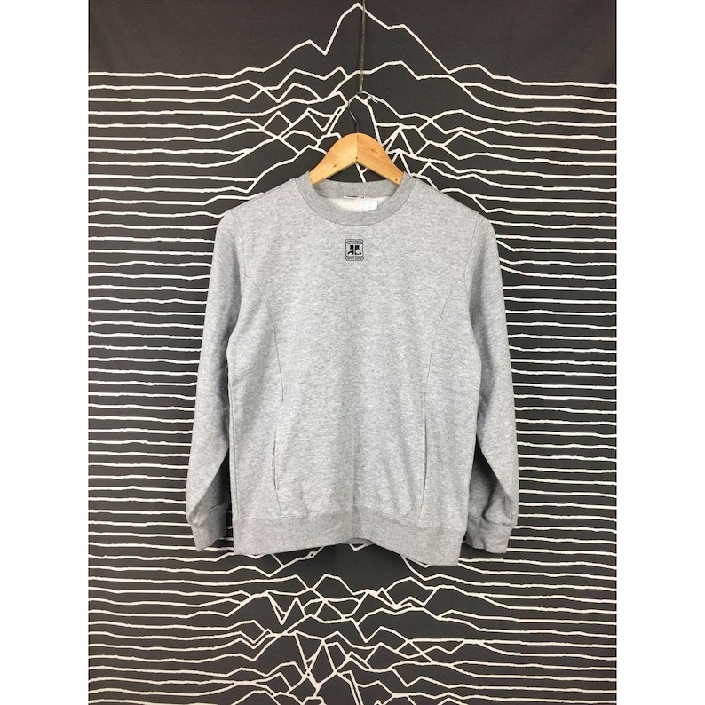 Peut inclure: Un sweat-shirt gris chin&eacute; &agrave; manches longues et col rond, suspendu &agrave; un cintre en bois. Le sweat-shirt pr&eacute;sente un petit logo carr&eacute; noir sur le haut de la poitrine gauche et des coutures verticales &agrave; l'avant.