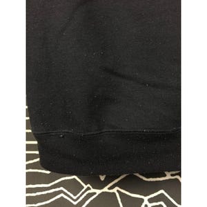 May include: Close-up of a black sweatshirt. The bottom hem and cuffs are a darker shade of black. The fabric has a slightly textured appearance, with small flecks visible on the surface. The background is a black and white patterned surface.