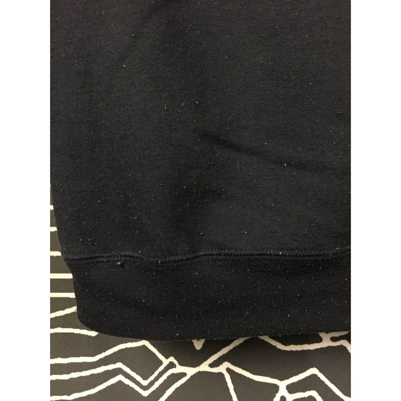 May include: Close-up of a black sweatshirt. The bottom hem and cuffs are a darker shade of black. The fabric has a slightly textured appearance, with small flecks visible on the surface. The background is a black and white patterned surface.