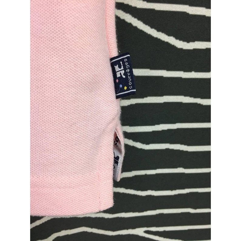 May include: Close-up of a light pink polo shirt. The shirt has a textured knit fabric and a small tag with the text "Courr&egrave;ges" in white on a navy blue background. The tag also features a logo with colorful dots.