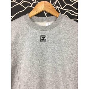 Peut inclure: Un sweat-shirt gris chin&eacute; avec un col rond et des manches longues, suspendu &agrave; un cintre en bois. Le sweat-shirt pr&eacute;sente un logo noir sur la poitrine avec le texte "courr&egrave;ges" et "sports futur".
