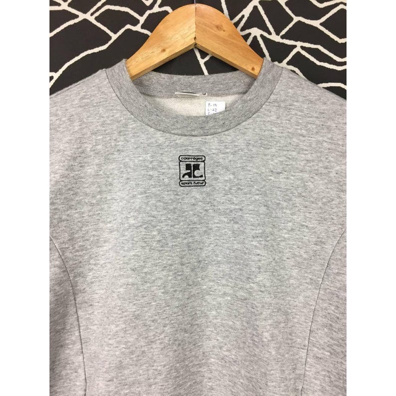 Peut inclure: Un sweat-shirt gris chin&eacute; avec un col rond et des manches longues, suspendu &agrave; un cintre en bois. Le sweat-shirt pr&eacute;sente un logo noir sur la poitrine avec le texte "courr&egrave;ges" et "sports futur".