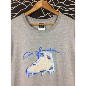 Peut inclure: T-shirt gris avec un graphisme bleu et blanc d'une basket montante et les mots "Air Jordan" en &eacute;criture bleue. Le design de la basket a des accents bleus et un effet de glace d&eacute;goulinante.