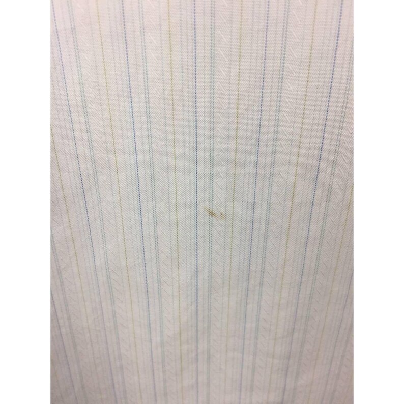 May include: A white fabric featuring a vertical pinstripe pattern in alternating light blue and pale yellow colors. The textile displays a subtle diagonal weave texture throughout the surface, with a small, faint brown spot visible near the center.