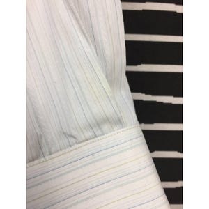 May include: A white long-sleeve shirt featuring a subtle vertical pinstripe pattern in light blue and olive green. The fabric has a fine, diagonal woven texture throughout, and the sleeve ends in a structured cuff with matching pinstripes.
