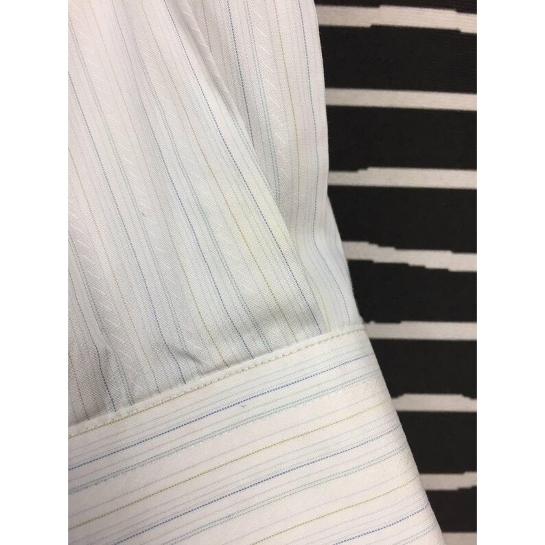 May include: A white long-sleeve shirt featuring a subtle vertical pinstripe pattern in light blue and olive green. The fabric has a fine, diagonal woven texture throughout, and the sleeve ends in a structured cuff with matching pinstripes.