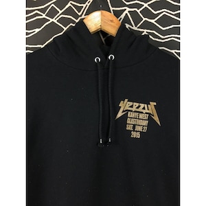 May include: Black hoodie with a drawstring hood. The front features a gold graphic that reads "YEEZY KANYE WEST GLASTONBURY SAT. JUNE 27 2015". The hoodie is made of a soft material.