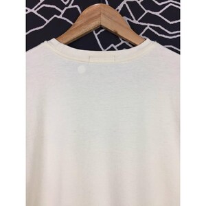 Op de afbeelding: Cr&egrave;mekleurig T-shirt aan een houten hanger. Het shirt heeft een ronde hals en een eenvoudig, minimalistisch ontwerp. De achtergrond is een zwart-wit geometrisch patroon.
