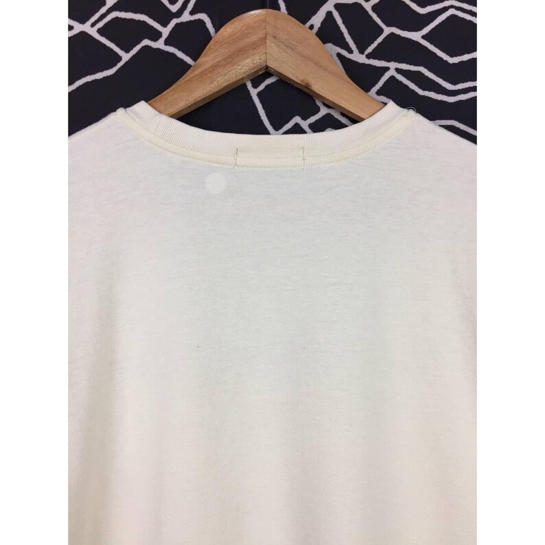 Op de afbeelding: Cr&egrave;mekleurig T-shirt aan een houten hanger. Het shirt heeft een ronde hals en een eenvoudig, minimalistisch ontwerp. De achtergrond is een zwart-wit geometrisch patroon.